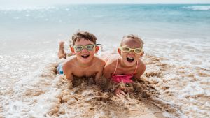 Siblings playing on beach, lying in water, having fun. Smilling girl and boy in swimsuits, swimming googles on sandy beach of Canary islands. Concept of family beach summer vacation with kids.