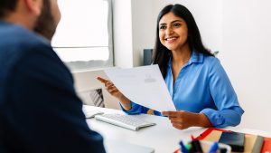 Confident successful Indian female HR recruitment manager holding resume in hands while having an interview in a modern office, looking and listening to job candidate. Friendly interview between HR female director and caucasian job candidate.