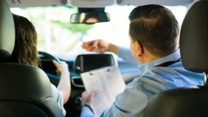Rear view of hispanic driving instructor guiding teen student during driving test lesson, holding clipboard and giving instructions