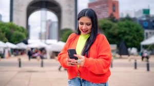 Smiling young Hispanic ethnic woman tourist taking using smartphone while standing in front of blurred Monument to the Revolution in Mexico City and saying hi with hand gesture in daylight