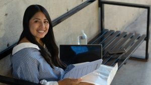 Young woman studying and working remotely outdoors on a modern bench, smiling at the camera while holding documents, a laptop and water bottle beside her