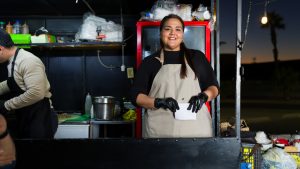Latina food truck worker wearing gloves, apron, taking customer orders with bright smile during evening shift